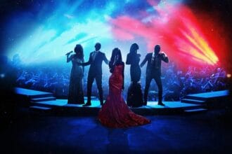 Five performers are silhouetted against an orchestra, they are dressed in formal wear and hold microphones. In the centre there is a woman who wears a red dress. Above the orchestra, the stage lights are red and blue